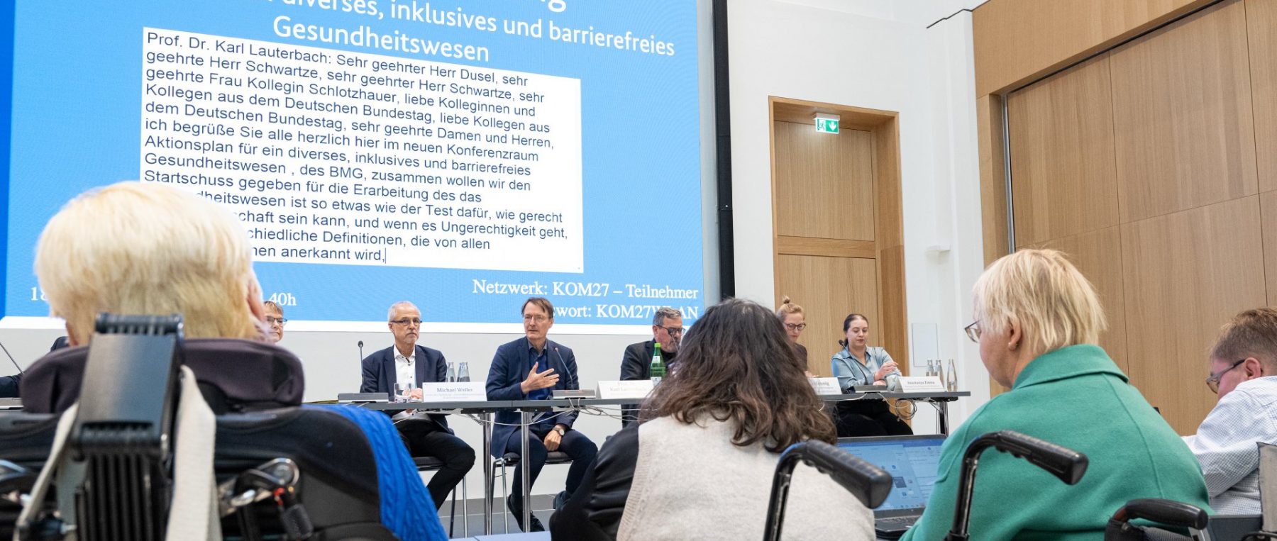 Menschen im Rollstuhl sitzen an Tischen dem Bundesminister für Gesundheit bei einem Treffen gegenüber. Im Hintergrund gibt es eine Beamerleinwand mit der Überschrift Auftaktveranstaltung