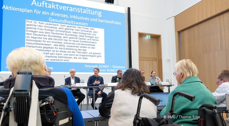 Menschen im Rollstuhl sitzen an Tischen dem Bundesminister für Gesundheit bei einem Treffen gegenüber. Im Hintergrund gibt es eine Beamerleinwand mit der Überschrift Auftaktveranstaltung