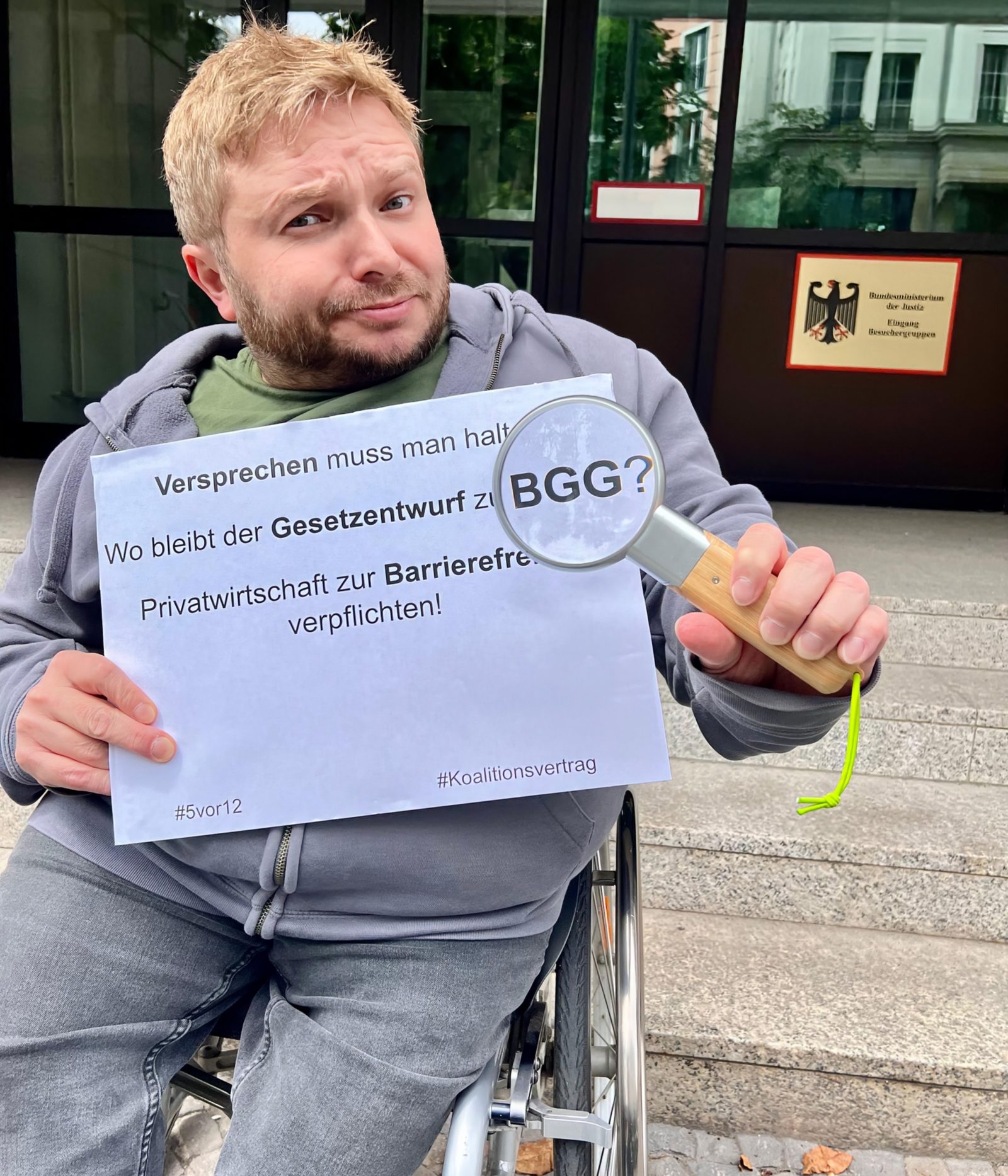 Alexander Ahrens steht mit seinem Rollstuhl vor der Treppe des Bundesjustizministeriums in Berlin und hält ein Schild hoch mit der Aufschrift: Versprechen muss man halten! Wo bleibt der Gesetzentwurf zum BGG? Privatwirtschaft zur Barrierefreiheit verpflichten! Dabei wird eine Lupe vor das Wort BGG gehalten, welches dadurch vergrößert wird. Foto: ISL e.V./Wiebke Schär Idee: Günter Heiden von Netzwerk Artikel 3