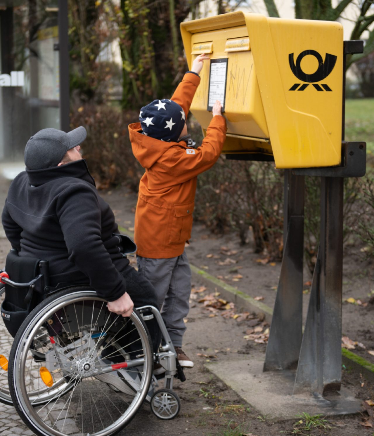 Mann im Rollstuhl steht mit Kind vor einem zu hohen Briefkasten