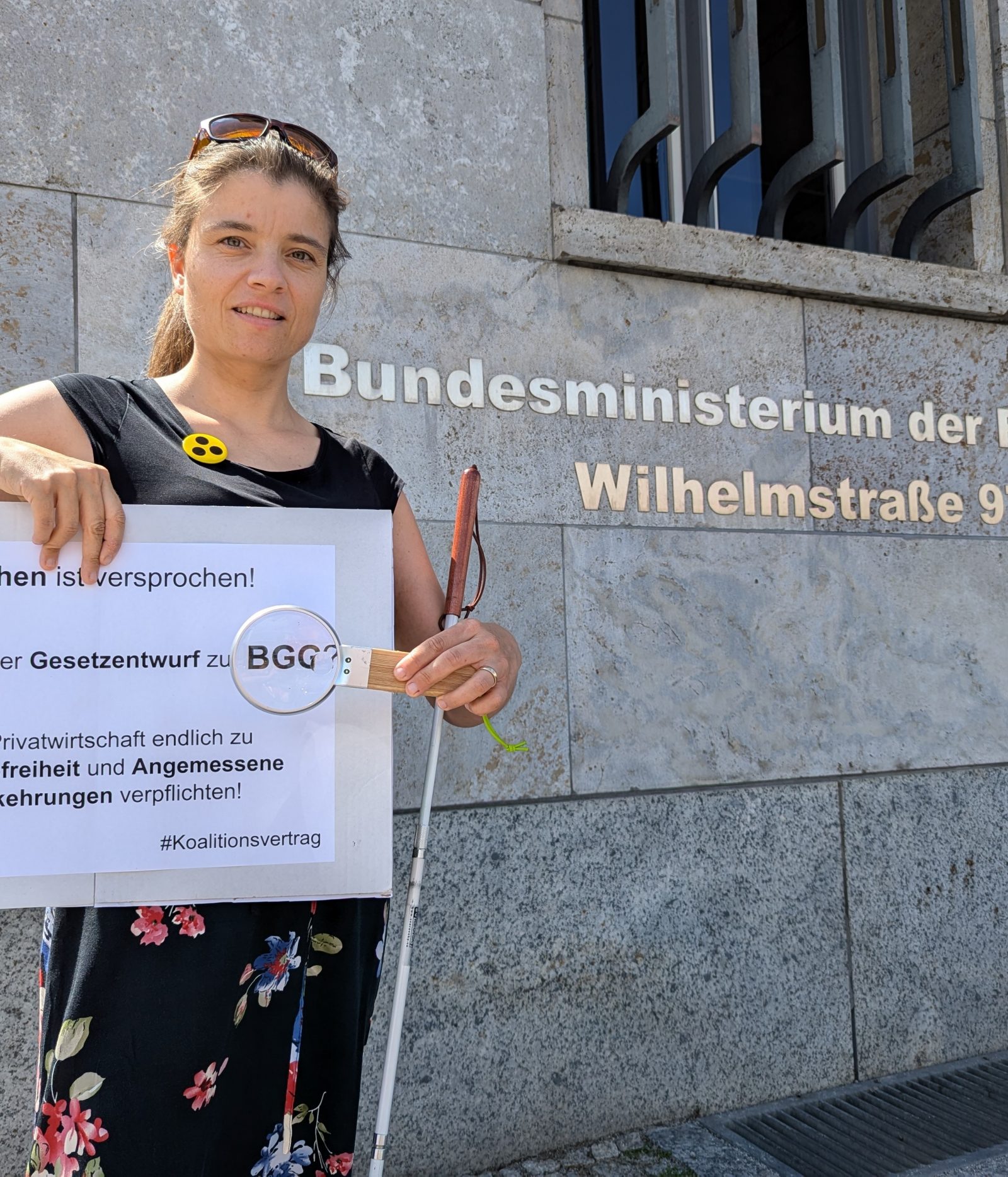 Bildbeschreibung: Juliane Harms steht vor dem Gebäude des Bundesfinanzministeriums in Berlin. Sie trägt einen Button mit Blindensymbol, hat ihren Langstock in der Armbeuge und hält eine Lupe vor dem Wort BGG auf dem Schild mit der Aufschrift „Versprochen ist versprochen! Wo steckt der Gesetzentwurf zum BGG? Die Privatwirtschaft endlich zu Barrierefreiheit und Angemessene Vorkehrungen verpflichten! #5vor12 #Koalitionsvertrag Foto: Alexander Ahrens ISL e.V. Idee: Günter Heiden Netzwerk Artikel 3 e.V.