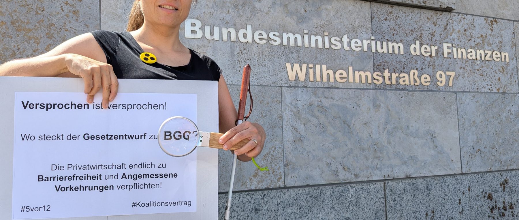 Bildbeschreibung: Juliane Harms steht vor dem Gebäude des Bundesfinanzministeriums in Berlin. Sie trägt einen Button mit Blindensymbol, hat ihren Langstock in der Armbeuge und hält eine Lupe vor dem Wort BGG auf dem Schild mit der Aufschrift „Versprochen ist versprochen! Wo steckt der Gesetzentwurf zum BGG? Die Privatwirtschaft endlich zu Barrierefreiheit und Angemessene Vorkehrungen verpflichten! #5vor12 #Koalitionsvertrag Foto: Alexander Ahrens ISL e.V. Idee: Günter Heiden Netzwerk Artikel 3 e.V.