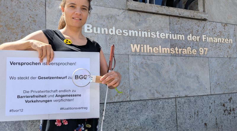 Bildbeschreibung: Juliane Harms steht vor dem Gebäude des Bundesfinanzministeriums in Berlin. Sie trägt einen Button mit Blindensymbol, hat ihren Langstock in der Armbeuge und hält eine Lupe vor dem Wort BGG auf dem Schild mit der Aufschrift „Versprochen ist versprochen! Wo steckt der Gesetzentwurf zum BGG? Die Privatwirtschaft endlich zu Barrierefreiheit und Angemessene Vorkehrungen verpflichten! #5vor12 #Koalitionsvertrag Foto: Alexander Ahrens ISL e.V. Idee: Günter Heiden Netzwerk Artikel 3 e.V.