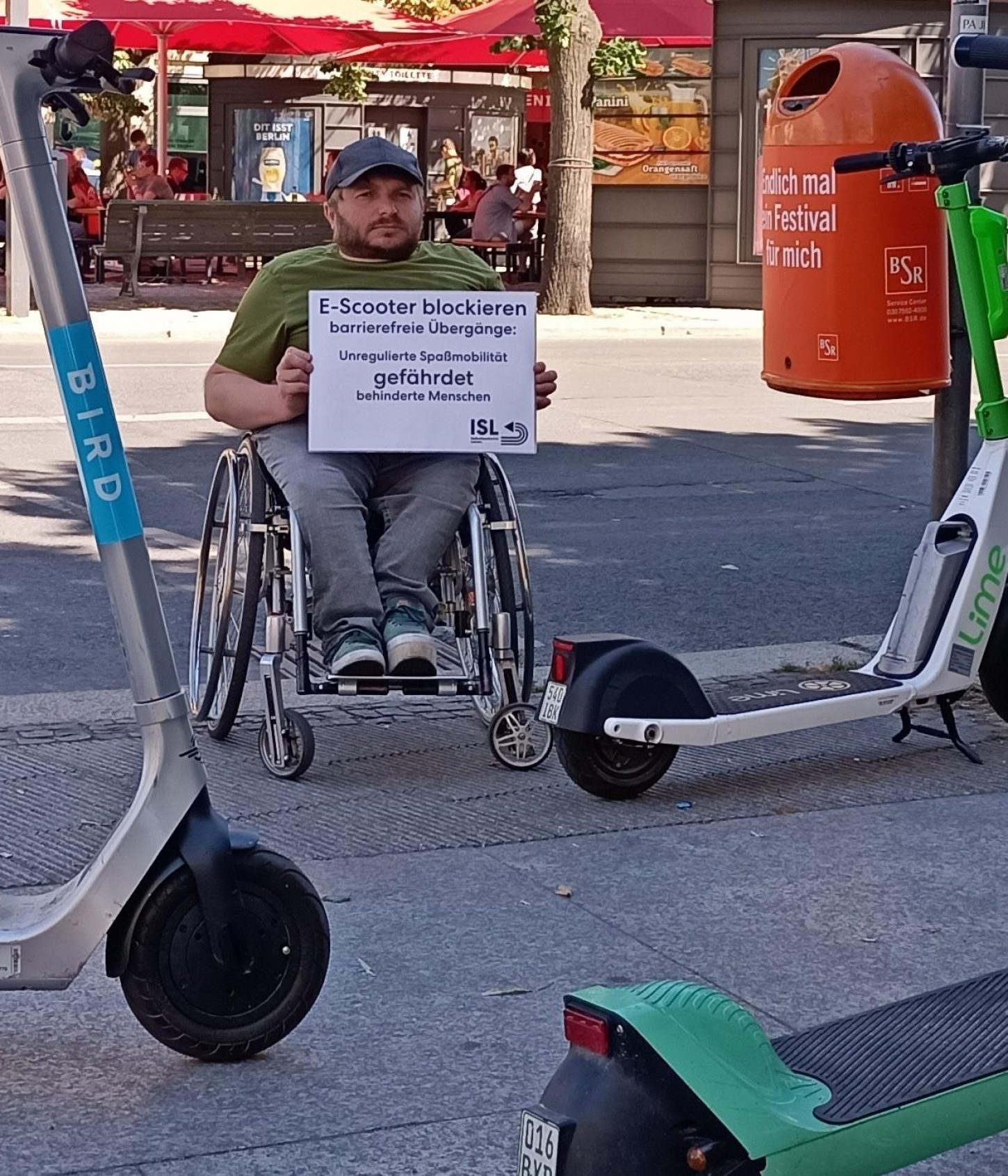 Mensch im Rollstuhl mit Schild steht an Ampelüberweg der mit E-Scooter zugestellt ist. Auf dem Schild steht: E-Scooter blockieren barrierefreie Übergänge. Unregulierte Spaßmobilität gefährdet behinderte Menschen. Foto: ABSV